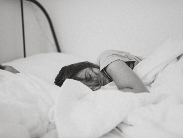 Mustavalkoinen kuva, jossa näkyy tummahiuksinen nainen nukkumassa valkoisessa sängyssä